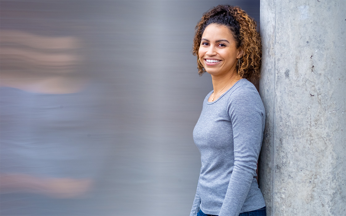 UW Tacoma alumna Saranda Ross leans against a cement pillar. She is wearing a grey shirt and has curly shoulder-length brown hair. To her left side is a blurry reflection of her that is meant to indicate motion.