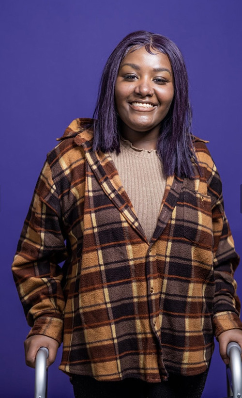 UW Tacoma student Chanise Jackson stands against a purple back drop. She is wearing a plaid shirt and his holding onto a walker.