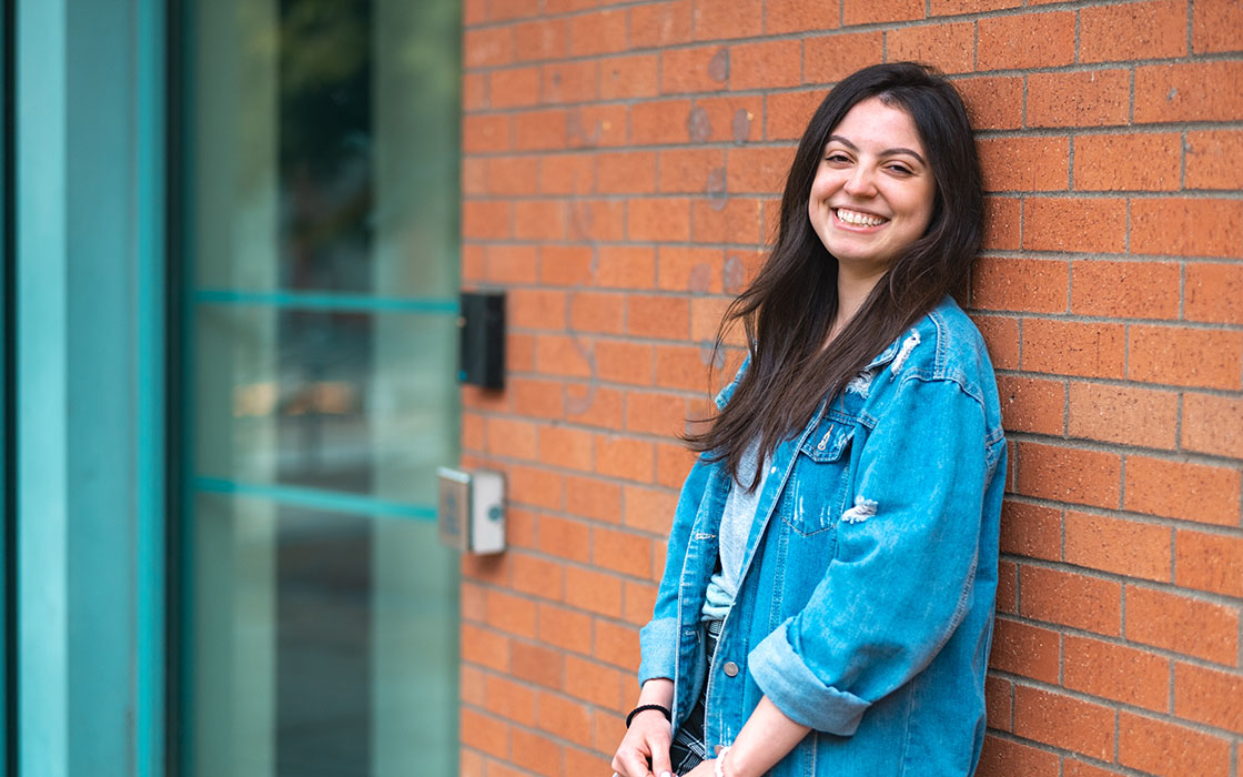 UW Tacoma senior Leticia Barreto leans against a red brick wall. She is wearing a blue denim jacket. Barreto has long black hair.