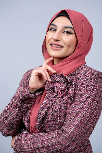 UW Tacoma student Maryam Al Darraji poses in front of a blank background. She is wearing a red headscarf and a plaid suit. Her left arm is folded across her stomach and her right arm is extended up with the index finger on her right hand touching her chin.
