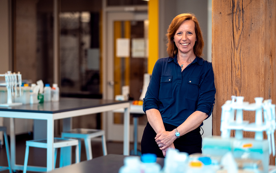 UW Tacoma faculty member Jutta Heller. Heller is leaning against a pillar. She is in a lab that has beakers and bottles in the foreground and on the background. Heller was red hair and his wearing a dark blue button up shirt.