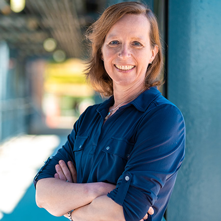 UW Tacoma faculty member Jutta Heller. Heller is leaning against a metal pole with her back against it. She has red hair and is wearing a blue button up shirt. Her arms are crossed at the waist.