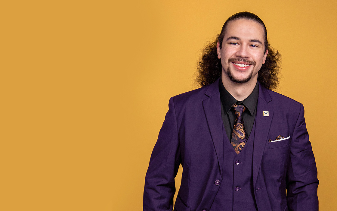 UW Tacoma student Jai'Shon Berry stands against a gold background. Berry is wearing a dark colored suit and has his hair pulled back in a ponytail. He also has a goatee. 