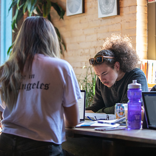 UW Tacoma student Jai'Shon Berry is sitting at a table studying. His head is turned down and he is looking at a book. Berry's hair is pulled back in a ponytail. A female student is in the foreground. She is wearing a white shirt and has long, dark hair.