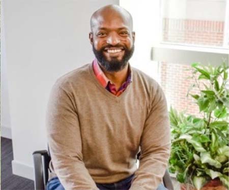 Davon Woodard in 2018, in tan sweater, with green plant on window ledge in background.