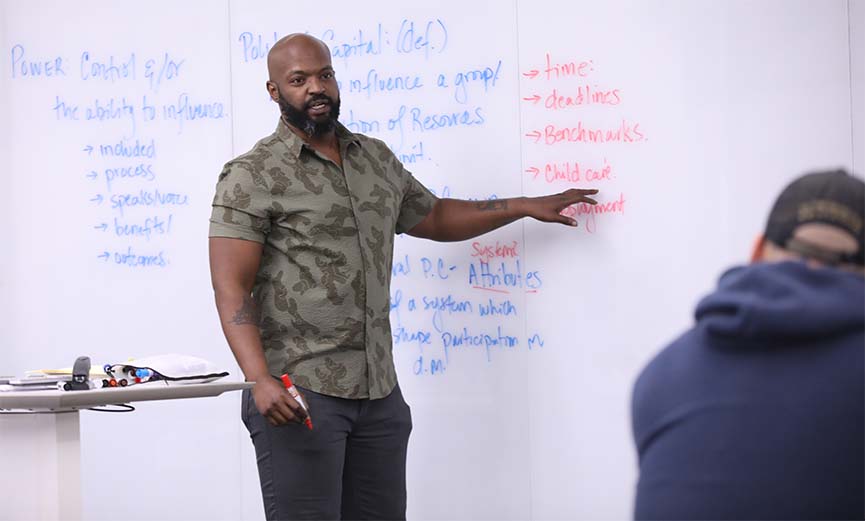 Davon Woodard pointing to whiteboard, holding red marker, looking toward student in blue hoodie and black cap.