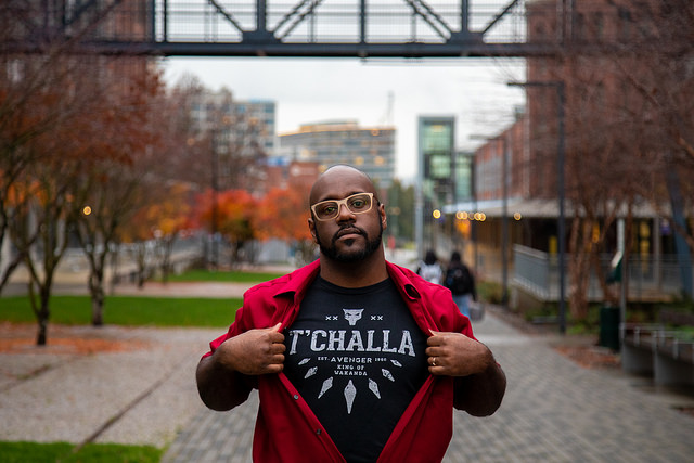 Dr. Chris Schell, standing on UW Tacoma Prairie Line Trail, pulling open shirt to reveal T'Challa/Wakanda t-shirt