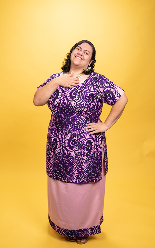 UW Tacoma student Exita Lealofi stands against a gold background. She is wearing multi-colored and patterned dress. Her right hand is held against her chest and her left hand is at her side. She has long, black hair.