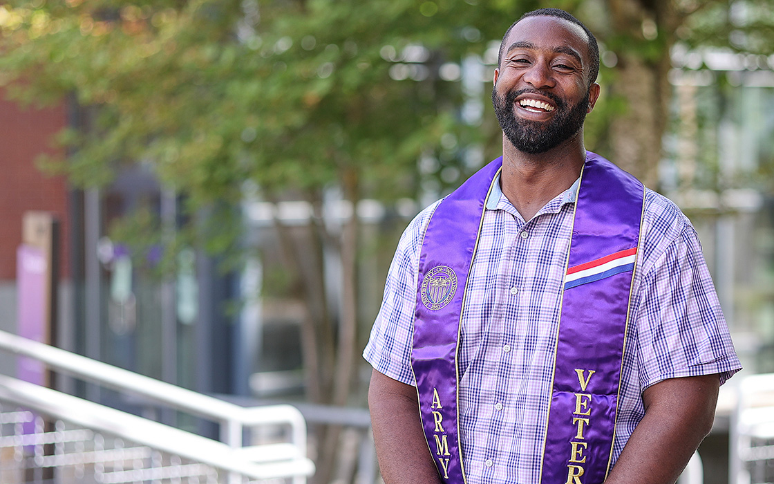 UW Tacoma alumnus Joe Davis stands in the right corner of the photo. He has short, black hair and is wearing a blue plaid button up with a purple stole around his neck that says veteran. In the background is a tree and an out of focus building.