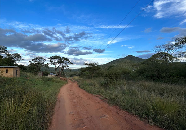 Picture of red dirt road on route to Vuu, with a cloud-speckled blue sky, lush greenery, and the Vuu mountain in the distance.