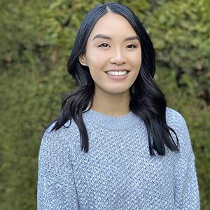 Headshot of Lan Allison. Allison stands in front of a bush with green leaves. She has long black hair and is wearing a purple patterned sweater.