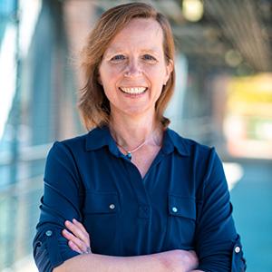 UW Tacoma faculty member Jutta Heller stands with her arms crossed across her mid-section. She has red hair and is wearing a dark blue button up shirt.