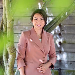   UW Tacoma faulty member Sunny Cheng stands underneath a tree. She has long black hair and is wearing a pink jacket. There is a wooden retaining wall in the background. Leaves from the tree can be seen in the front of the photo.