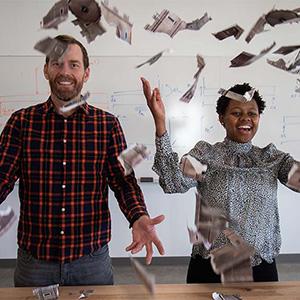 A man and a woman stand in front of a white board. They have their hands outstretched. There are pieces of grey colored carboard floating in the air around them. These are pieces of a torn up cardboard castle.