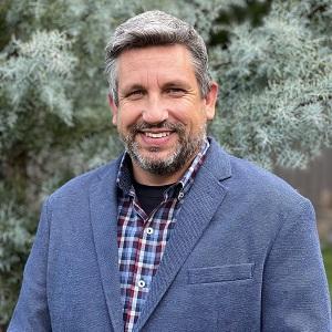 Grey-haired white man in a blazer and plaid shirt smiles with trees behind him