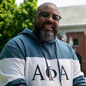 Headshot of UW Tacoma alumnus Jeff Dade. Dade is wearing a green and white sweatshirt with Greek (fraternity) letters on it. He is also wearing dark colored glasses. In the background is a tree with green leaves and the red bricked Snoqualmie Powerhouse building.