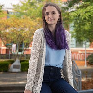 UW Tacoma student Holly Wetzel sits on a railing on campus. There are trees in the background and you can see part of the Snoqualmie Library. Wetzel is wearing a long white coat, blue jeans and a white shirt. Her hair is brown with purple tips.