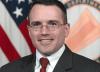 Christopher Martinez, wearing a suit and tie in front of an American flag