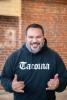 A Chamorro man with beard in a Black hoodie that says Tacoma in front of a red brick wall.