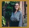 a photo of Gabriela Romero. She has glasses and short brown hair. She is wearing a blue blouse and standing in front of a plant covered brick wall.