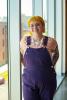 Kat Key, a student employee, stands inside by large glass windows, wearing purple overalls and a striped shirt.