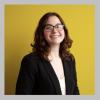 a photo of Moira Kelley. She has shoulder length curly brown hair and brown glasses. She is wearing a black blazer and standing in front of a gold background.